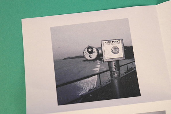 A black and white photograph of a coin-operated telescope overlooking a coastline. A sign on the viewer reads “VIEW POINT”.