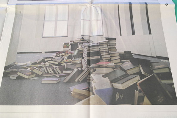 A large pile of books is scattered across a grey floor in a bright room. Some books are stacked in a precarious tower, while others lie haphazardly around the base.