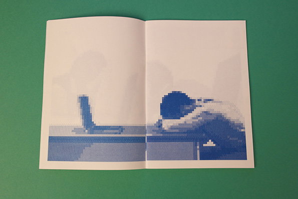 An open booklet shows a blue and white pixelated image across two pages. A figure is seen from the side, leaning over a surface next to a laptop.