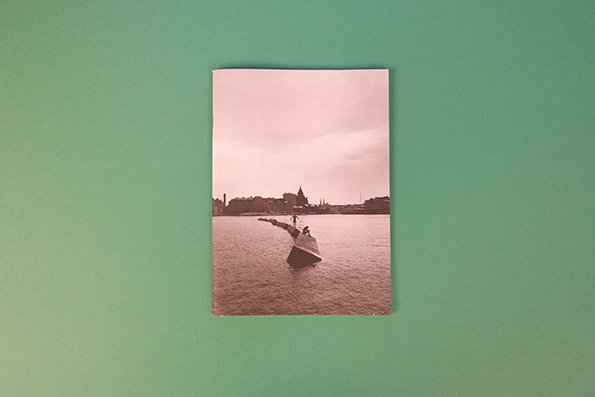A faded, sepia-toned photograph rests on a plain green surface. It shows a person standing on a concrete pier that extends into a body of water towards a distant city skyline.