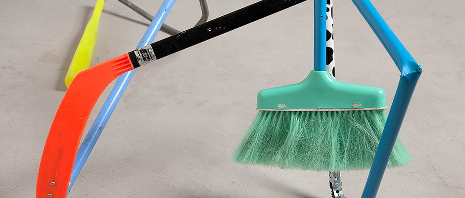 A black and orange hockey stick leans against a blue metal frame. Beside it, a green broom with a black and white patterned handle rests on a grey floor.