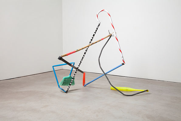 A sculpture made from a chaotic assembly of everyday objects in a white gallery space. The structure includes a blue plastic frame, a green broom head, a red and white striped pole, and a long grey cable.