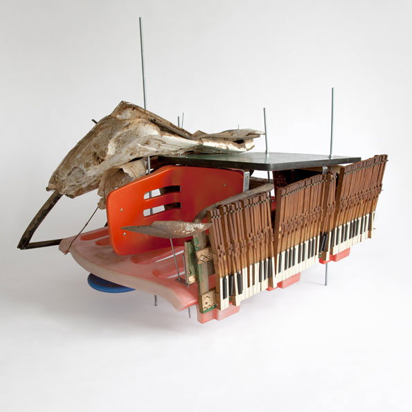 A sculpture made from a mix of found objects, including a red plastic chair and a keyboard. A large, jagged piece of pale stone or concrete rests on top, held in place by thin metal rods.