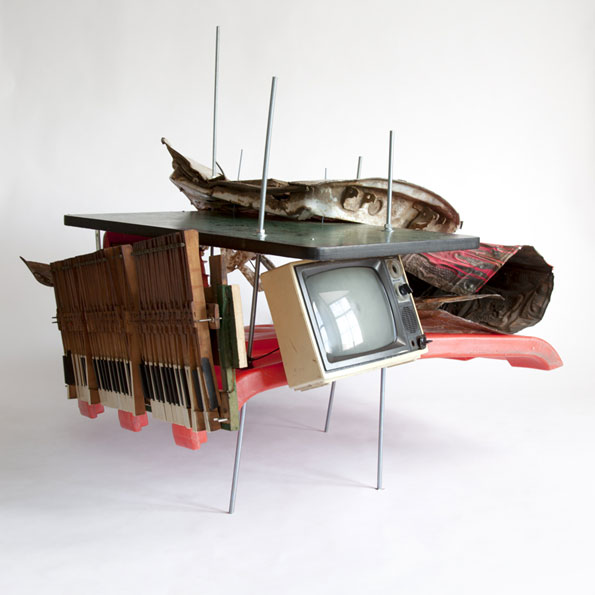 A sculpture made from a chaotic assembly of found objects, including a vintage television, a keyboard, and a red plastic chair. The items are held together by thin metal rods and a dark flat board against a plain white background.