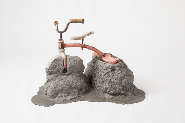 A sculpture of a bicycle partially submerged in two mounds of grey, textured concrete. Only the handlebars, a small section of the frame and the seat remain visible above the surface.
