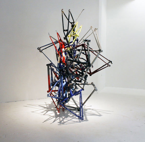 A sculpture made from a tangled heap of bicycle frames in red, blue, yellow and silver. The frames are welded together into a jagged, vertical spire in a white gallery space.