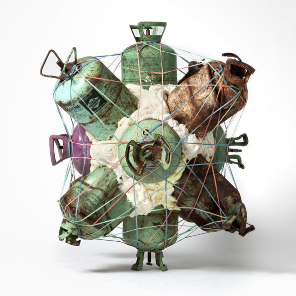 A sculpture of several weathered gas canisters arranged in a spherical cluster. The canisters are held together by a network of thin blue and red wires, surrounding a central mass of white, textured material.