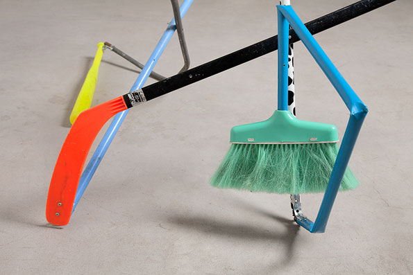A collection of sports equipment and tools lean against each other on a grey floor. An orange hockey stick crosses over a light blue frame and a green broom.