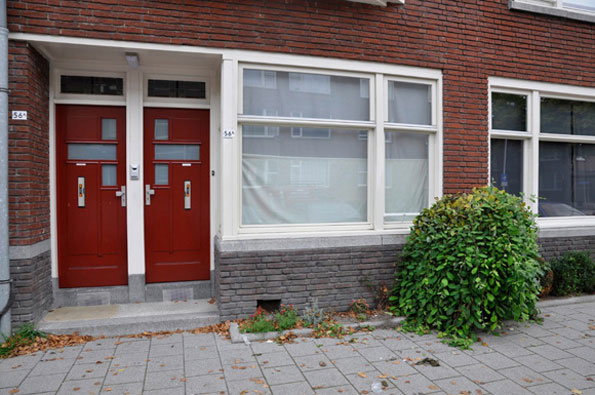 Two red doors stand side by side in the entrance of a red brick building. To the right, a large window is partially covered by a white blind, with a green shrub growing beside it.