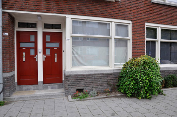 Two red doors stand side by side in the entrance of a red brick building. A large window with white frames and closed blinds sits next to the doors, beside a rounded green shrub.