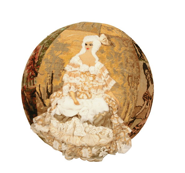 A circular textile work featuring a figure in an ornate, cream-coloured 18th-century gown. The figure is rendered in a mix of embroidery and fabric, set against a golden, textured background.