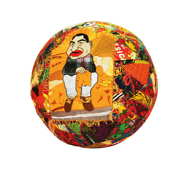A textile collage on a circular canvas featuring a stylised figure with red lips sitting in a chair. The figure is surrounded by a vibrant mix of patterned fabrics in red, yellow and orange.