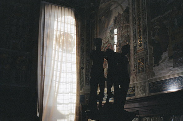 Three silhouetted figures stand in a dimly lit room beside a large window with sheer white curtains. Ornate paintings and gold detailing cover the surrounding walls.