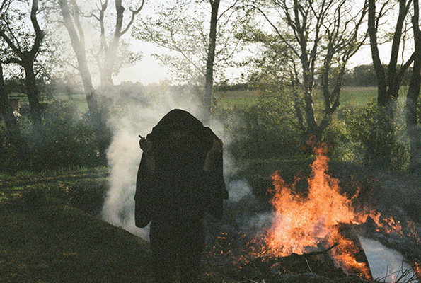 A person draped in a heavy black cloak stands beside a roaring bonfire in a wooded field. Thick white smoke swirls around them against a backdrop of thin, leafless trees.