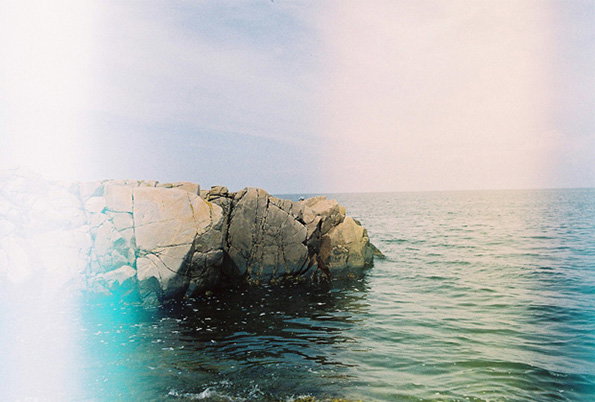 Large, jagged grey rocks sit in a calm sea under a pale sky. A bright, blurred wash of white and cyan light obscures the left side of the frame.