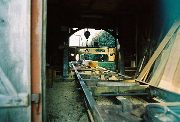 A view from inside a dark wooden shed looking out towards a bright green landscape. A long metal conveyor belt runs through the centre, carrying heavy stone blocks.