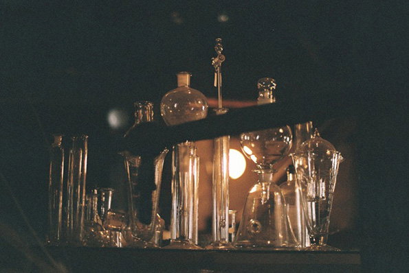 A collection of clear glass vessels and laboratory flasks are arranged on a dark surface. A person’s arm reaches across the frame to adjust one of the objects.