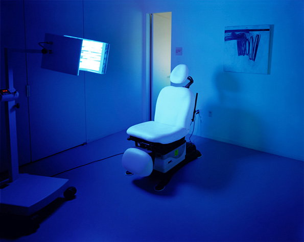 A white medical chair sits in a room bathed in deep blue light. To the left, a glowing screen is mounted on a stand, and a small abstract painting hangs on the far wall.