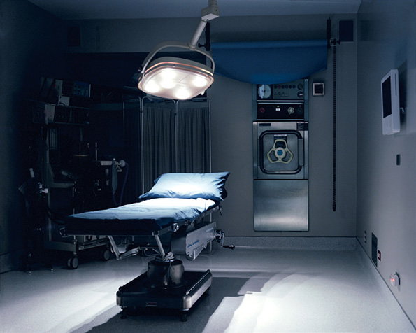An empty operating theatre with a blue surgical bed positioned under a bright overhead light. Medical equipment and a stainless steel unit stand against the grey walls in the dim room.