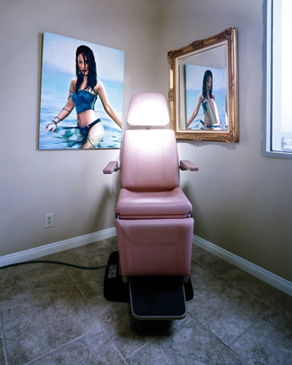 A pink medical examination chair sits in the centre of a plain room. Behind it, a painting and a gold-framed mirror both show a person in a blue outfit standing in water.