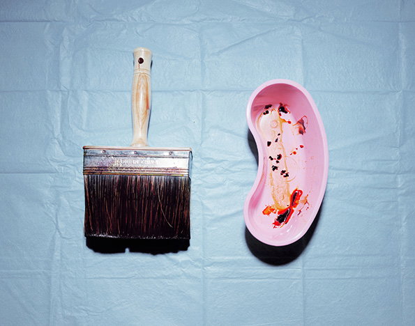 A wide paintbrush with dark bristles and a wooden handle sits beside a pink, kidney-shaped medical tray. The tray contains smears of red and orange paint against a light background.