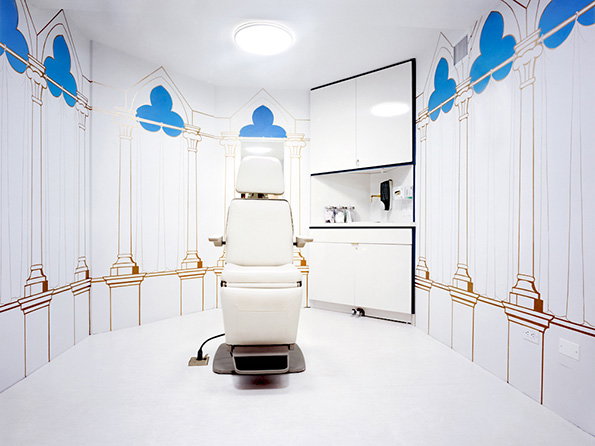 A white medical treatment chair sits in the centre of a bright, all-white room. The walls are decorated with blue and gold line drawings of gothic arches and columns.