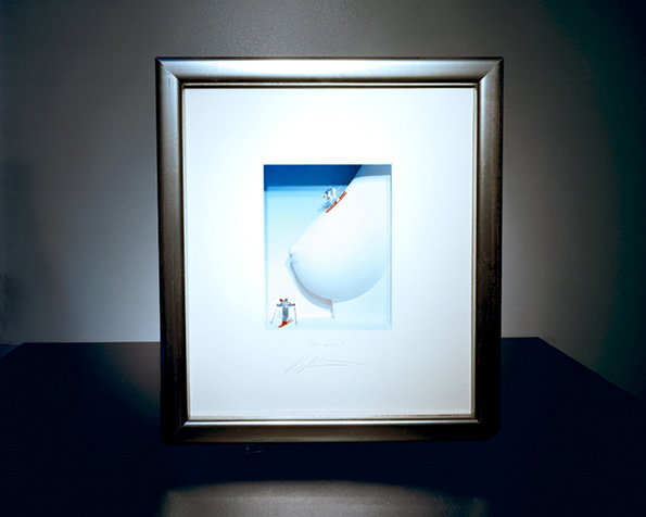 A framed 3D artwork features a large, white, rounded form resembling a breast. Two tiny figures stand on the surface, one at the peak and one at the base.