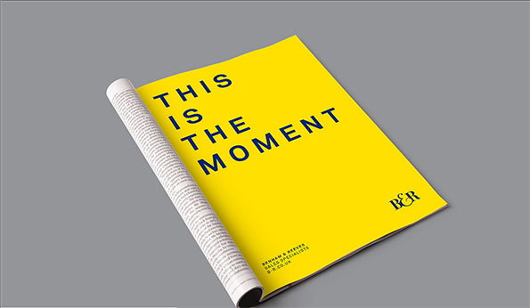 A bright yellow brochure lies on a grey surface, with its pages partially curled. Bold black text reads “THIS IS THE MOMENT”, with a small “B&R” logo in the corner.