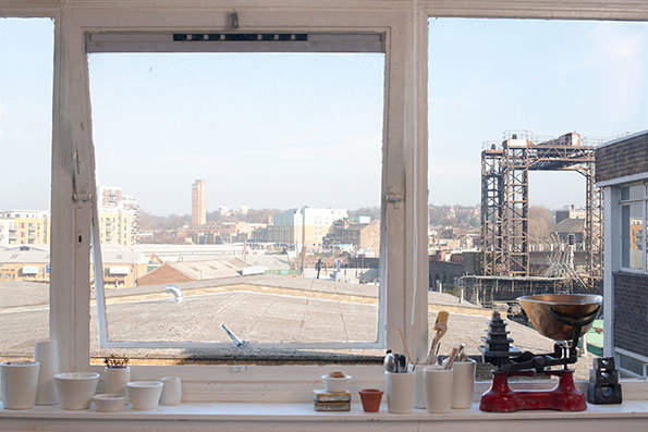 A view through an open white window frame overlooking a city skyline. On the windowsill sit several small white ceramic pots and a red vintage kitchen scale.
