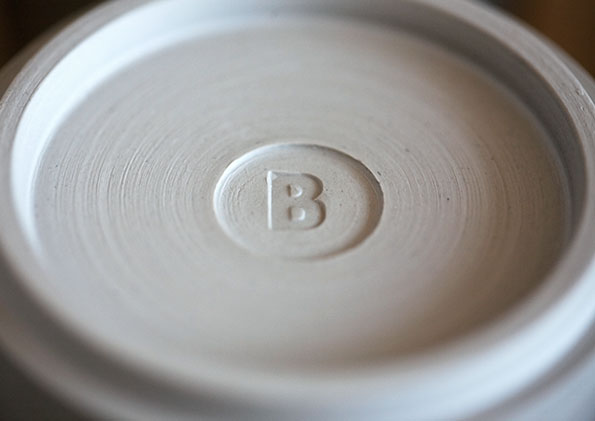 A close-up of the base of a white ceramic object. A capital letter “B” is embossed within a small circular indentation in the centre.