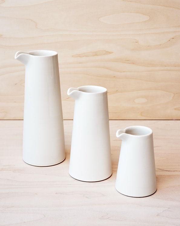 Three white ceramic jugs of varying heights stand in a row against a light wood background. Each jug has a tapered cylindrical shape and a small pouring lip.