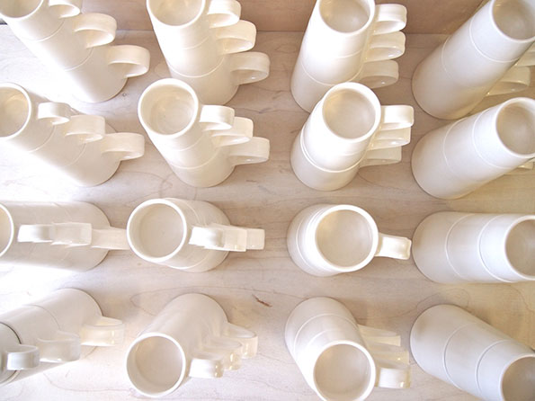 Rows of white ceramic mugs are arranged in a light-coloured wooden tray. Many of the mugs are stacked inside one another, with their handles aligned.