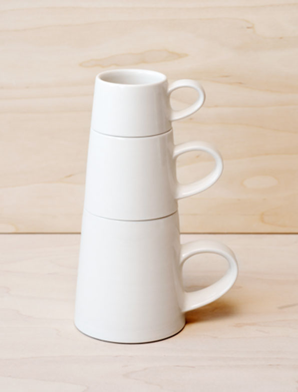 Three white ceramic mugs of increasing size are stacked on a light wood surface. The mugs have a tapered, conical shape and curved handles.