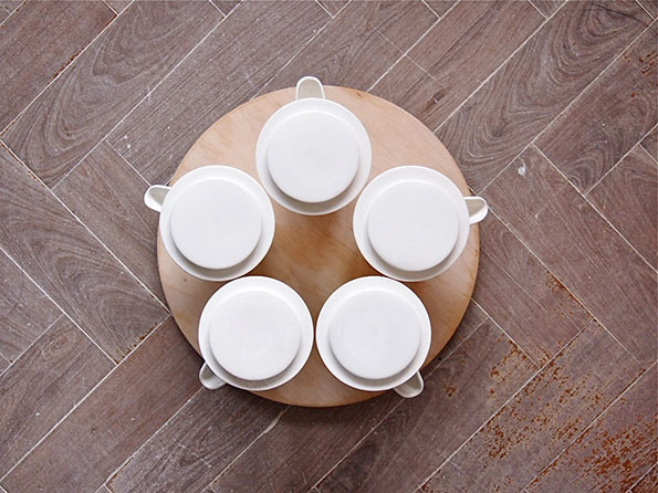 Five white ceramic pots with lids are arranged in a circle on a round wooden board. The board sits on a brown wooden floor with a herringbone pattern.