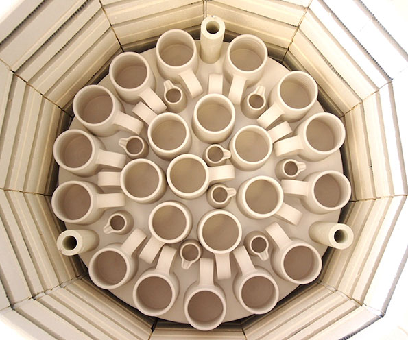 A top-down view of dozens of unglazed ceramic mugs and small cups arranged closely together. They are packed into a circular kiln, ready for firing.