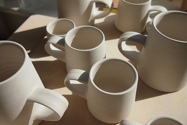 A collection of unglazed ceramic mugs in various sizes sit on a light wooden workbench. The pale clay pieces are arranged in a cluster, catching bright, direct sunlight.
