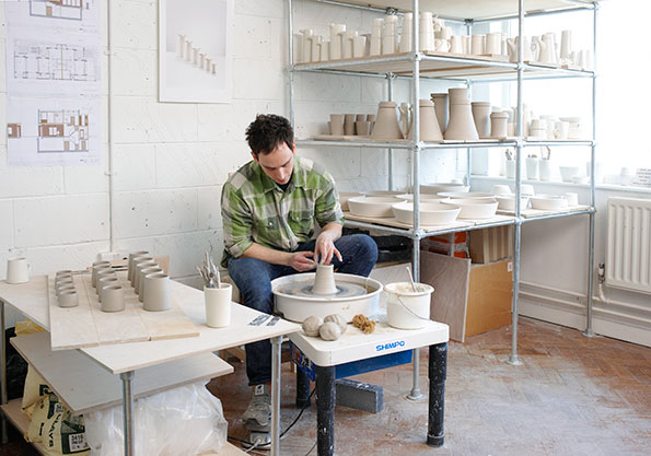 A person shapes a piece of clay on a pottery wheel in a bright studio. Behind them, metal shelves are filled with rows of white ceramic vessels in various sizes.