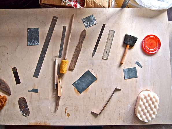 A collection of printmaking tools and materials are laid out on a light wood surface. The items include brayers, metal rulers, a small mallet, and several ink slabs.