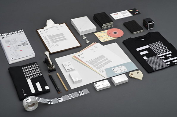 A brand identity kit laid out on a grey surface. The collection includes a black T-shirt, notebooks, business cards, a clipboard and a roll of branded tape.