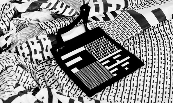 A black tote bag with a white geometric pattern rests on a pile of fabric. The surrounding material features matching black and white abstract prints.