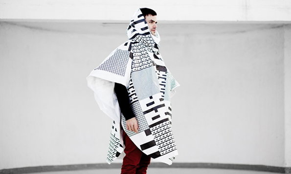 A person stands in profile against a plain white wall, wrapped in a large black and white patterned fabric. The fabric features various geometric grids and abstract shapes, draped over their head and shoulders like a cloak.