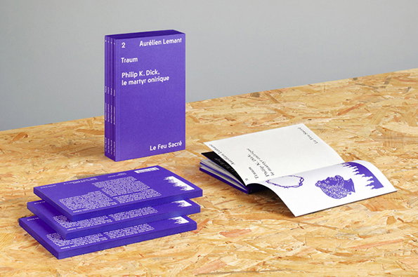 A purple book stands upright on a wooden surface, with a stack of purple pamphlets and an open booklet beside it. The book spine reads “2 Aurélien Lemant”, “Traum”, “Phillip K. Dick, le martyr onirique” and “Le Feu Sacré”.