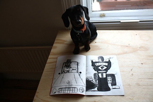 A black dachshund with a blue collar sits on a light wood table. In front of them lies an open sketchbook featuring two black and white illustrations, one of which includes the word “REBELS”.