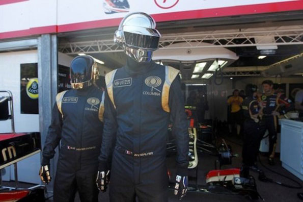 Two figures stand in a racing garage wearing black racing suits and mirrored helmets. Their suits feature the “Lotus” and “Columbia” logos.