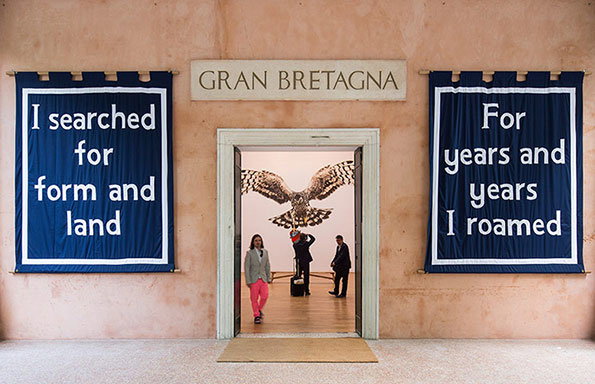 Three people stand in the doorway of a building labelled “Gran Bretagna”. Above them, a large image of a bird of prey with outstretched wings is displayed on the interior wall. Two blue banners flank the entrance, reading “I searched for form and land” and “For years and years I roamed”.