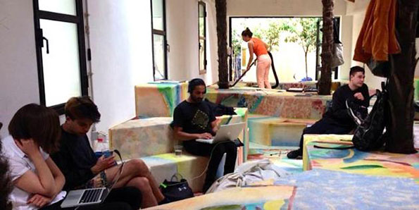 Four people relax in a bright room filled with large, colourful foam blocks. Three people sit on the blocks using laptops and phones, while a fourth person vacuums the floor in the background.
