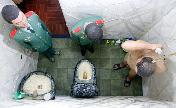 A high-angle shot of three figures in a small, white-tiled room with two toilets. Two figures wear green military uniforms, while a third person is shirtless and wearing blue flip-flops.