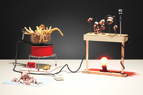 A sculpture of a deep fryer contains fried octopus tentacles, sitting atop a red pot. To the right, raw octopus pieces are skewered on a wooden stand above a glowing red light bulb.