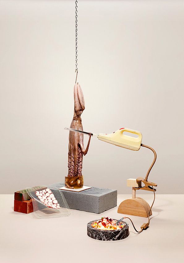 A large squid hangs from a metal chain, suspended above a grey concrete block. A handheld power saw is positioned as if cutting through the squid's body.