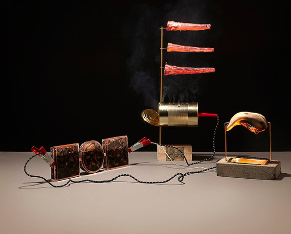 A sculpture featuring a series of connected objects on a grey surface against a black background. Three pieces of raw meat are skewered on a vertical stand above an open tin can, with steam rising from the can. To the left, three square frames are linked by wires and clips, while a piece of meat rests on a stand over a heated block to the right.